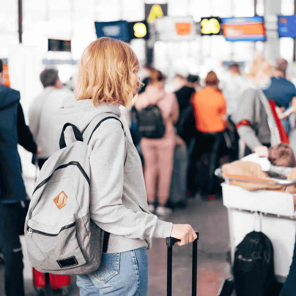 A traveller with a backpack and carry-on luggage standing in an airport check-in area, surrounded by other passengers.