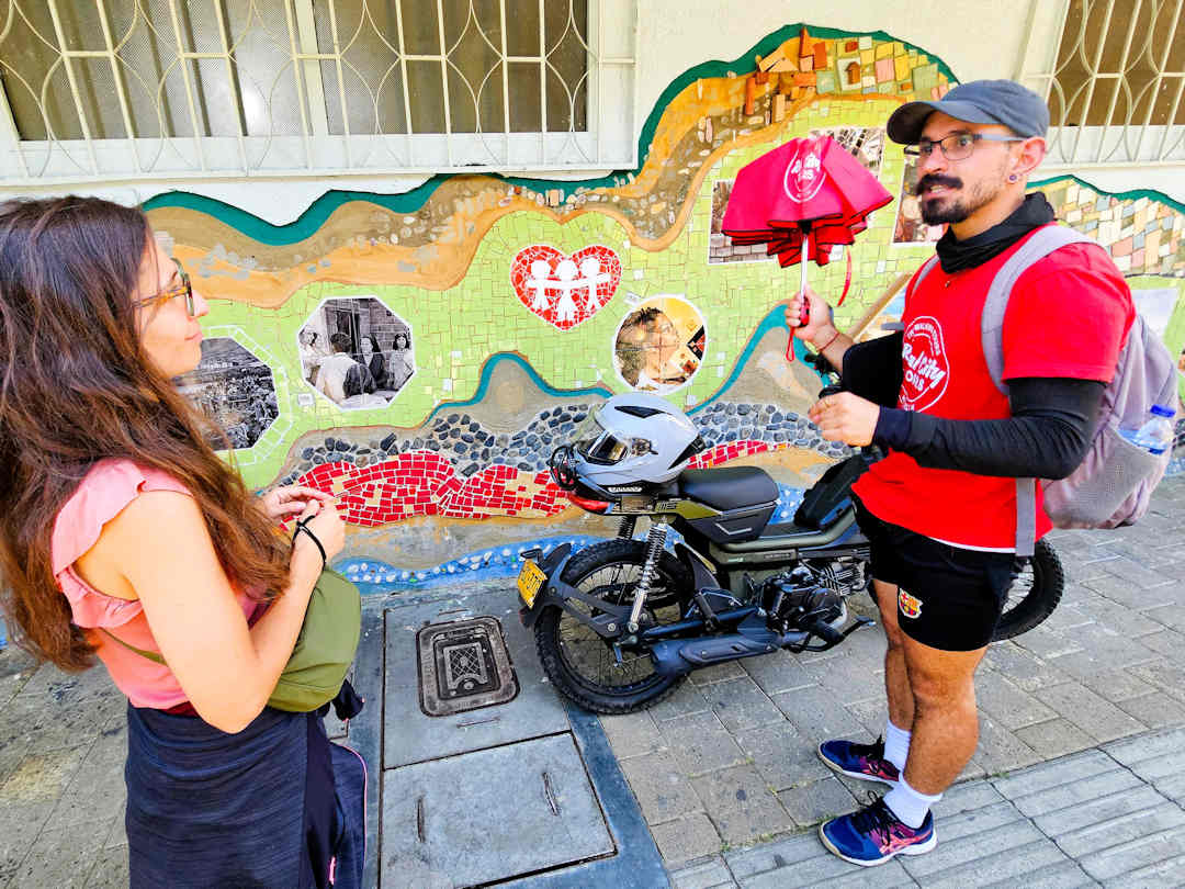 guide toto with tour participant outside el bosque school Guide explaining a colourful mosaic mural in Moravia during the Comuna 4 Barrio Transformation Tour, Medellín