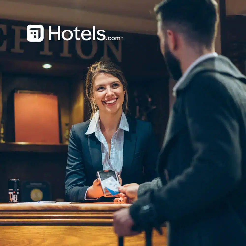Guest checking into a hotel reception, handing a phone to a smiling receptionist, with the Hotels.com logo visible above.
