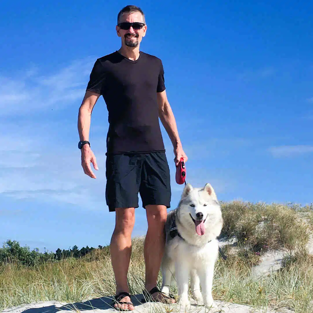 Siberian Husky named Mia standing on a beach next to Paul, both enjoying a sunny day.