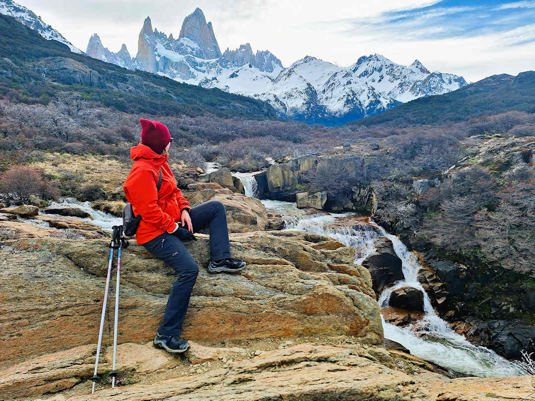 Sandra wearing a Patagonia Women's Torrentshell 3L Rain Jacket resting by a waterfall on the Fitz Roy River with Mount Fitz Roy in the background near El Chaltén, Argentina