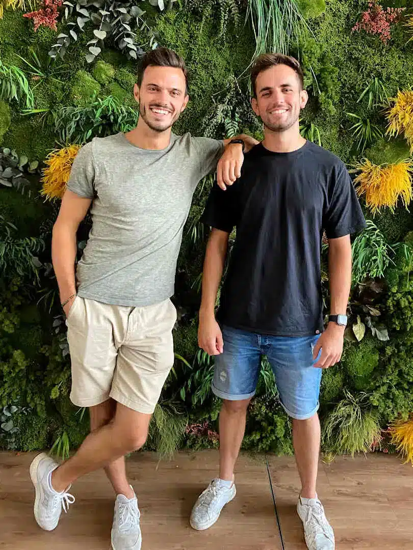 Alexander Haufschild and Nils Lohmann, casually dressed, stand smiling in front of a vibrant green wall of plants. They are the co-founders of Socialbnb, a platform for sustainable tourism.